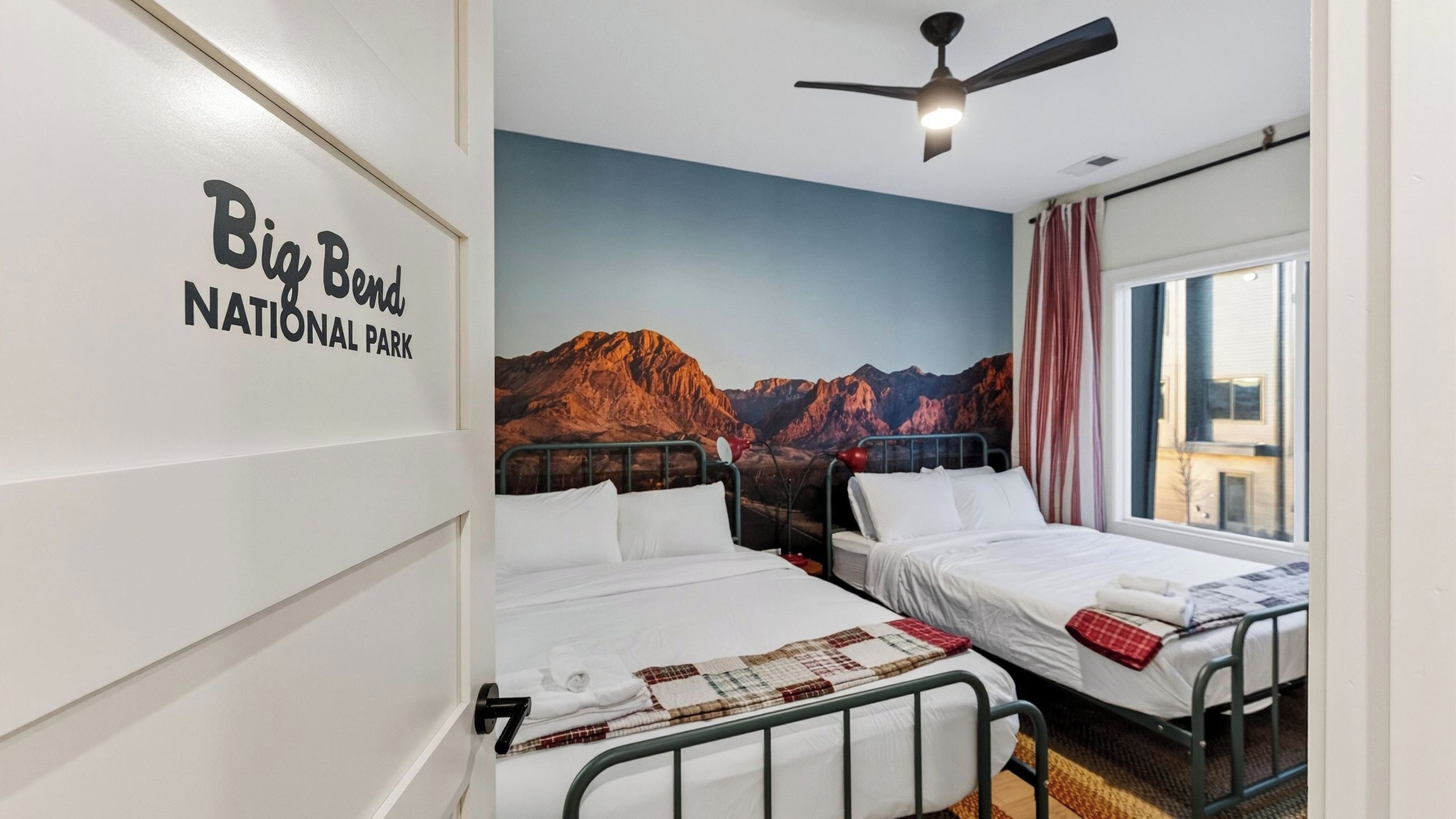 Ein Hotelzimmer mit zwei Betten, einem Deckenventilator, einem Wandbild von Bergen und einem Fenster mit Vorhängen.