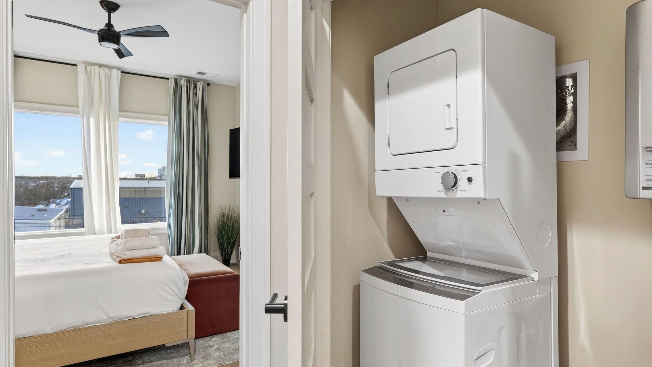 A hotel room with a bed, a ceiling fan, a washing machine, and a window with curtains.