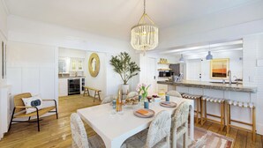 A modern kitchen with white cabinetry, a dining area with a white table and wicker chairs, and a hanging chandelier. - Venice Veranda by Avantstay 3-min Walk to Beach + Rare Walk Street EV Charger + Firepit + Outdoor Dining (Venice)