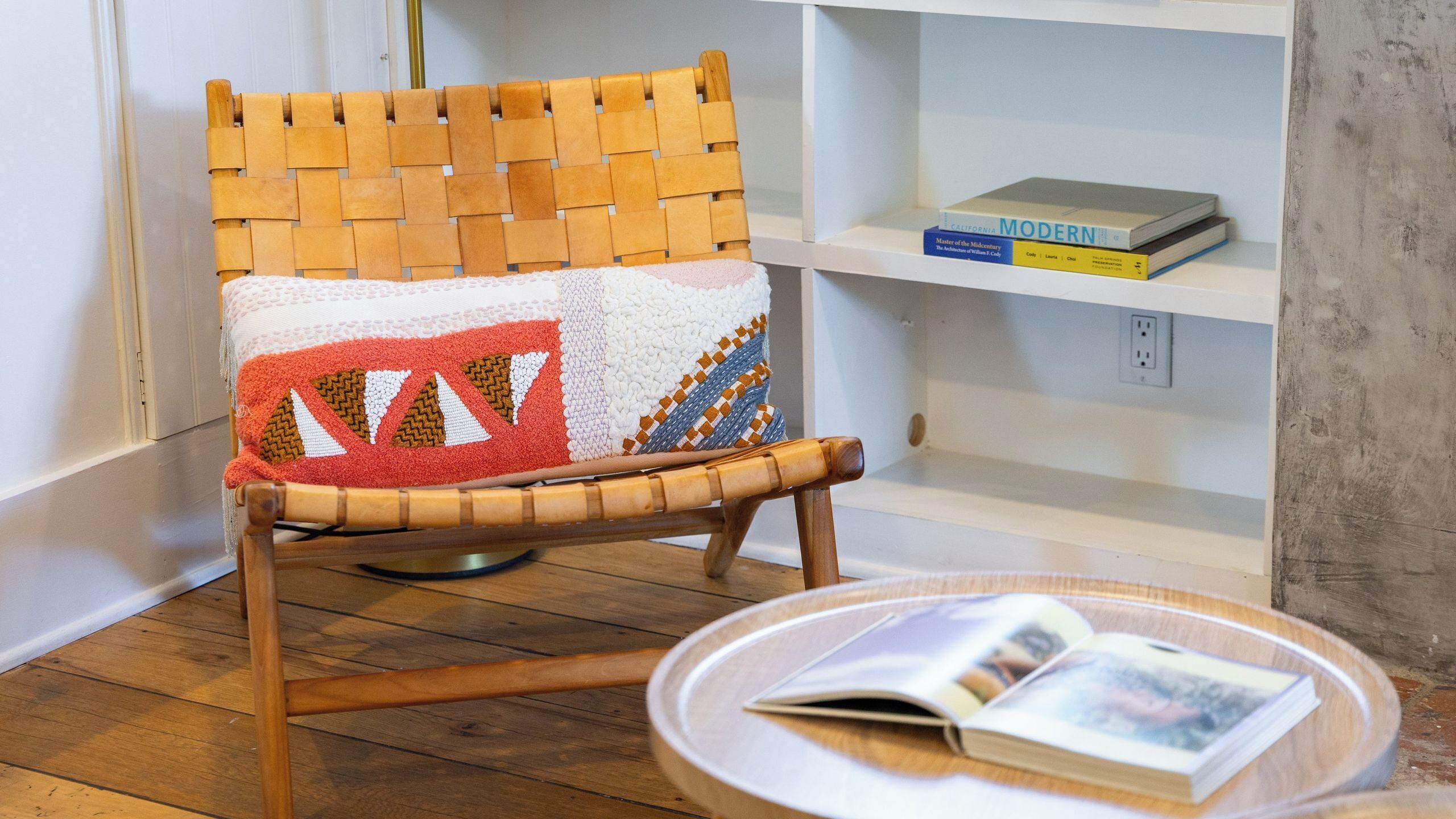 A wooden chair with a woven backrest and a cushion featuring a geometric pattern. A white shelf with books and a round table with a magazine are also visible.