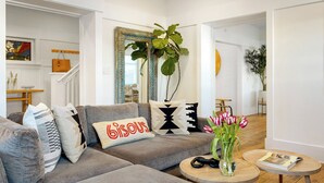 A living room with a grey sofa, patterned cushions, a mirror with a decorative frame, a plant, and a vase with flowers. - Venice Veranda by Avantstay 3-min Walk to Beach + Rare Walk Street EV Charger + Firepit + Outdoor Dining (Venice)