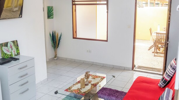 A room with a red sofa, a white dresser, a glass coffee table, and a colorful rug. There is a window with blinds, a door leading outside, and a small plant on the floor. - Appartement Standard a Cayenne Baduel (Cayenne)