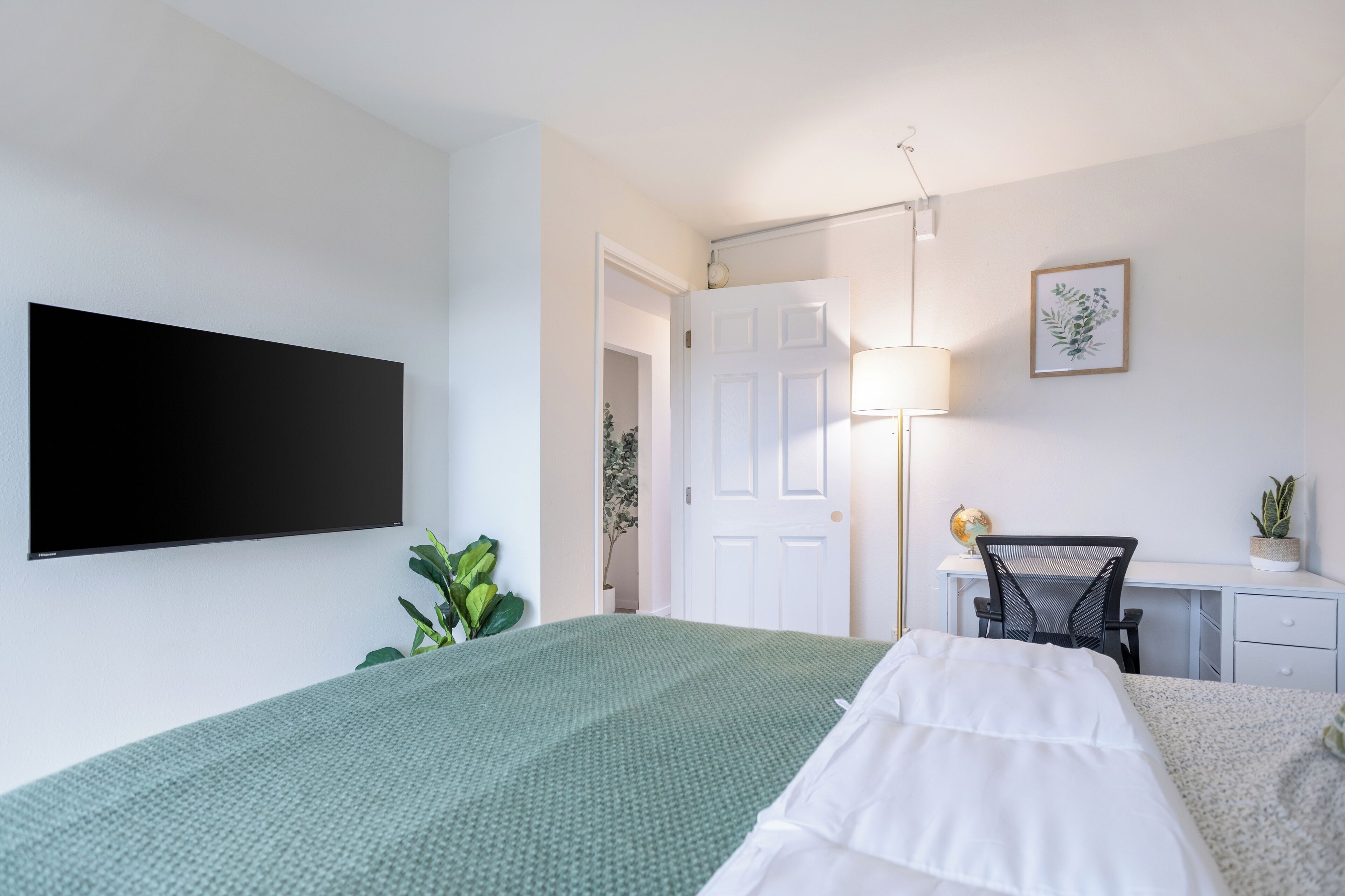 A modern hotel room with a large bed, a desk with a chair, a flat-screen TV mounted on the wall, a lamp, and a small potted plant.