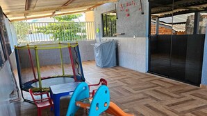 Un espace extérieur abrité doté d’un plancher en bois, d’une structure de jeu bleue et jaune, et d’une chaise rouge.