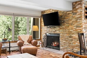 A living room with a stone fireplace, a flat-screen TV mounted above it, a comfortable leather armchair, and a large window offering a view of greenery. - Spacious Vail Golf & Ski Condo, Pool, Hot Tub (Vail)
