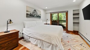 Ein Schlafzimmer mit einem Bett, einem Schreibtisch mit Lampe, einem Gemälde, einem Fenster mit Blick und einem an der Wand befestigten Fernseher.