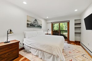 Ein Schlafzimmer mit einem Bett, einem Schreibtisch mit Lampe, einem Gemälde, einem Fenster mit Blick und einem an der Wand befestigten Fernseher.