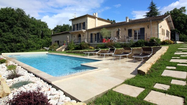 Ein großer Pool mit Liegestühlen und einem gepflasterten Weg, der zu einem Haus mit Ziegeldach führt.