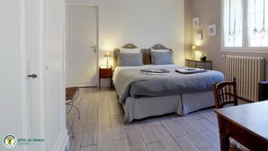 Ein Schlafzimmer mit einem Doppelbett, Nachttischen, einem Sessel, einem Tisch und einem Fenster mit Vorhängen.