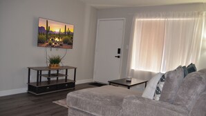Un salón con un sofá gris, una mesita de centro, un televisor montado en la pared y una puerta con cortina blanca.