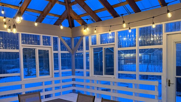 Un porche cubierto con mesa y sillas de madera, ventanas amplias y luces colgantes.