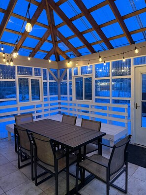 A covered patio with a wooden table and chairs, large windows, and string lights. - Dandy House E-bikes/Sauna (Homer)