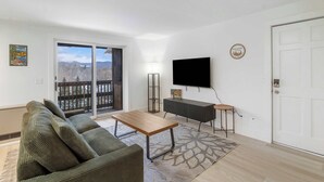 En stue med en sofa, et sofabord, et fladskærms-tv, et lille sengebord og en dør.