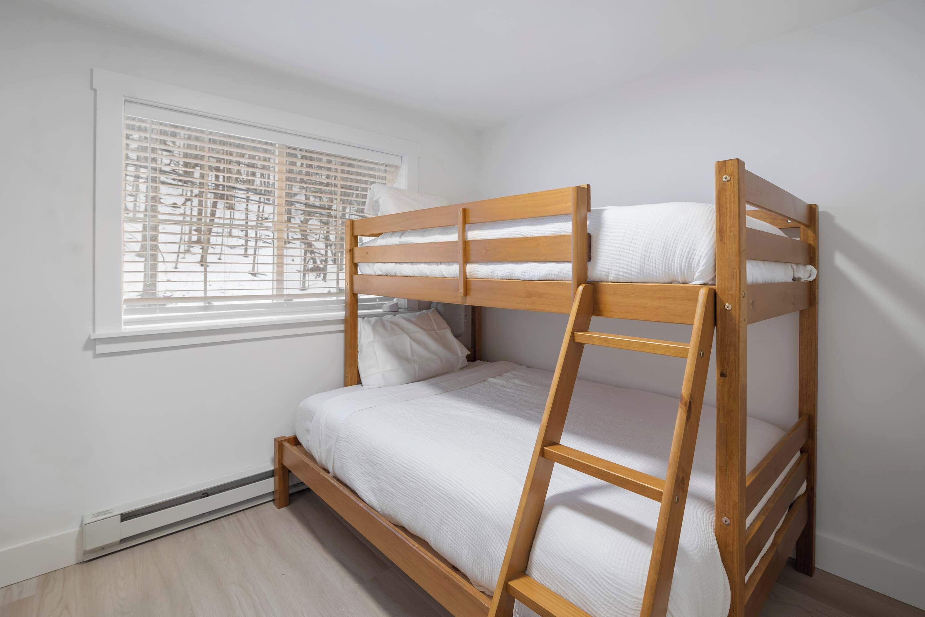 Een stapelbed met houten frame en wit beddengoed in een kamer met raam en radiator.