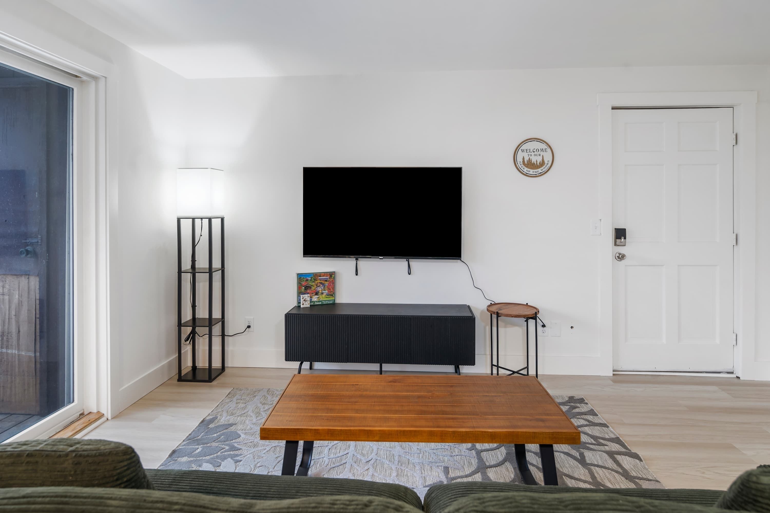 Ein Wohnzimmer mit Couchtisch aus Holz, einer schwarzen Unterhaltungsecke mit Fernseher darüber, einem kleinen Beistelltisch und einer Tür mit einer Willkommensschild.