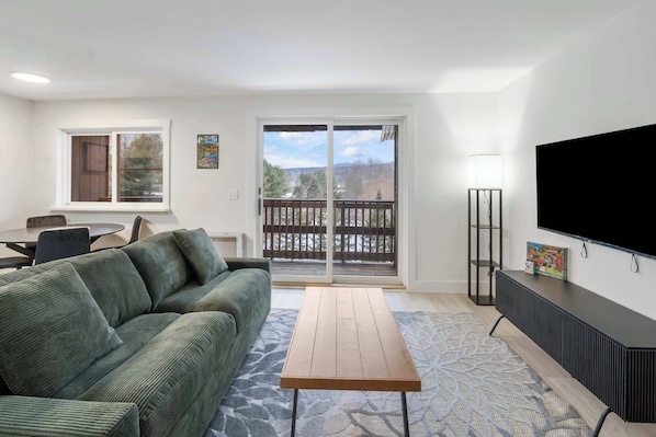 A living room with a green sofa, a wooden coffee table, a TV on the wall, and a balcony with a view of trees and a fence.