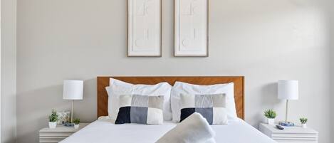 A neatly made bed with a rolled white towel, two bedside tables with lamps, and a wall with two framed abstract art pieces.