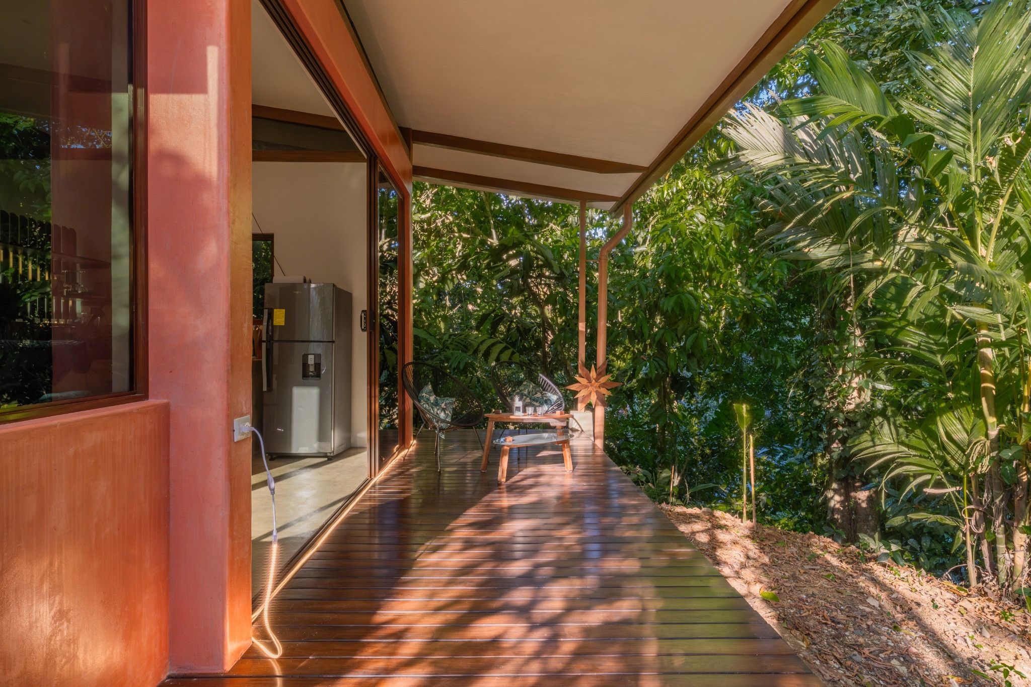 A covered wooden deck with a refrigerator and a chair, surrounded by lush greenery.