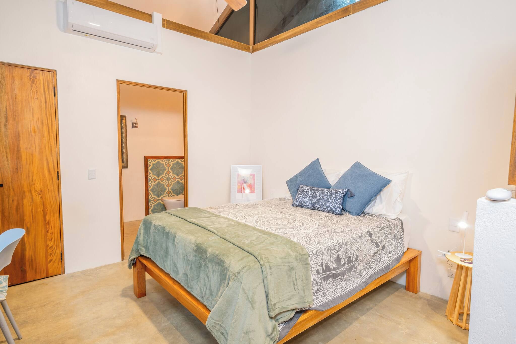 A bedroom with a bed, a chair, a bedside table, and an air conditioning unit.