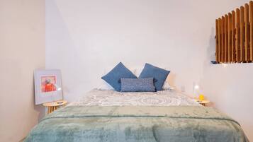 A bedroom with a bed, pillows, a blanket, a bedside table, a lamp, and a wall-mounted light fixture.
