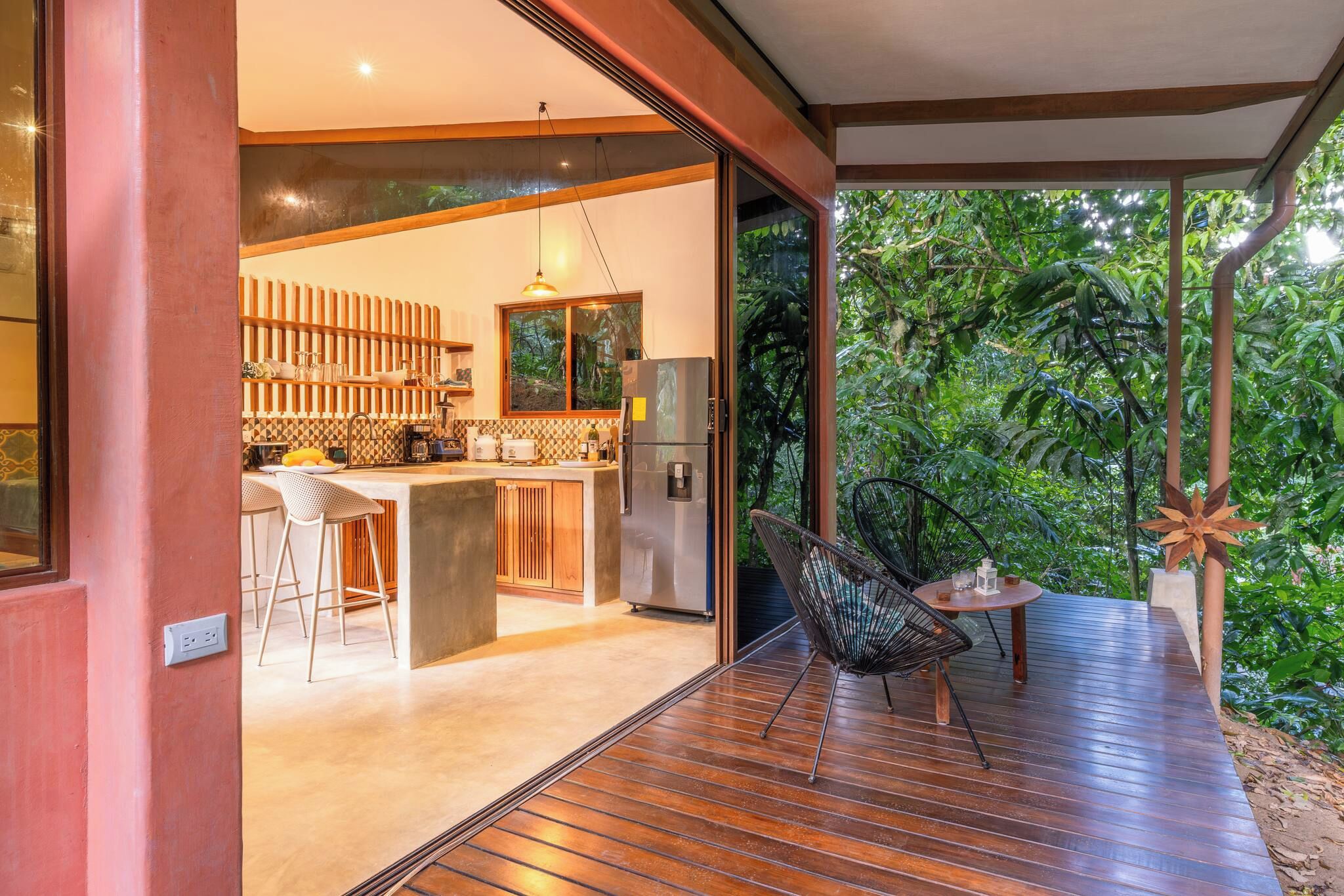 A modern kitchen with a bar area, a dining table with chairs, and a view of lush greenery.