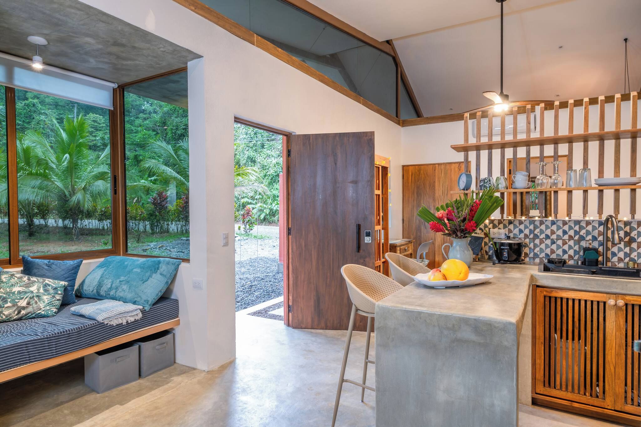 A modern kitchen with a bar area, a seating area with a view of greenery, and a large wooden door.