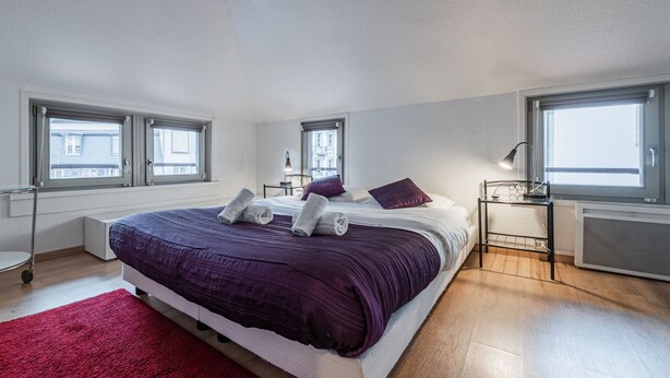 Ein Schlafzimmer mit einem großen Bett, zwei Fenstern, einem Heizkörper und einem roten Teppich.