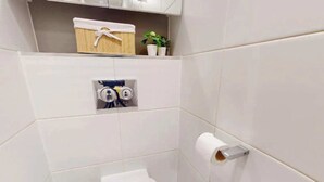 Ein kleines, modernes Badezimmer mit weißen Fliesen, einer Toilette, einem Handtuchhalter und einem Regal mit Pflanze und Korb.