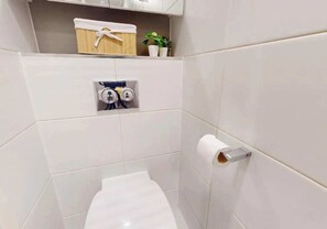 A small, modern bathroom with white tiles, a toilet, a towel holder, and a shelf with a plant and a basket. - Student Only Paris Gardens (London)