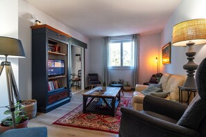 A living room with a sofa, armchair, coffee table, bookshelf, and a TV. - L'Appartement du Belon - Proche GR34 (Moëlan-sur-Mer)
