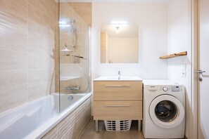 Een moderne badkamer met een douchecabine van glas, een witte wastafel met spiegel erboven, een houten wastafelmeubel met lades, en een voorlader wasmachine.