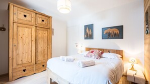 Een slaapkamer met een houten garderobe, een bed met wit beddengoed, twee aan de muur bevestigde schilderijen en een nachtkastje met een lamp.