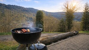 Un barbecue avec des saucisses et de la viande, installé devant un paysage montagneux, avec des arbres et un ciel dégagé.