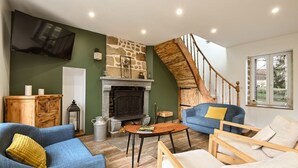 A living room with a stone fireplace, a wooden staircase, and a flat-screen TV mounted on the wall. - Le Lotus - 1000 m² de jardin (Putanges-le-Lac)