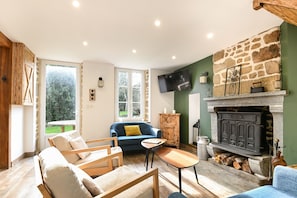 A cozy living room with a stone fireplace, a blue sofa, wooden furniture, and a television mounted on the wall. - Le Lotus - 1000 m² de jardin (Putanges-le-Lac)