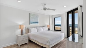 Une chambre à coucher comprenant un lit, des tables de chevet, un ventilateur de plafond et une grande fenêtre donnant sur la plage.