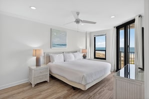 Ein Schlafzimmer mit einem Bett, Nachttischen, einem Deckenventilator und einem großen Fenster mit Blick auf den Strand.