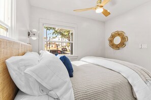 Une chambre à coucher avec un lit, un ventilateur de plafond, une fenêtre donnant sur des immeubles et un miroir décoratif.