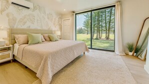 A bedroom with a large bed, a nightstand, a wall-mounted air conditioner, a window with a view of trees, and a floor-to-ceiling mirror. - Spacious 4-bedroom condo in gorgeous Kahuku with AC (Kahuku)