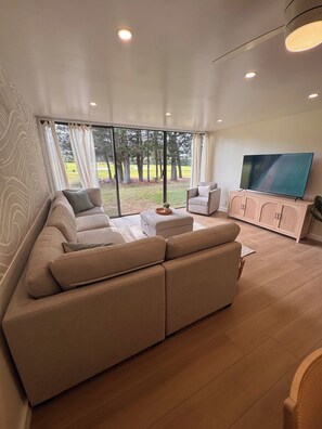 A modern living room with a large sectional sofa, a television on a wooden cabinet, and a view of a landscaped area through large windows. - Royal Turtle Palms: Luxe 4BR Turtle Bay Condo, Sleeps 12 (Kahuku)