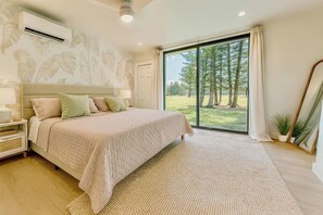 A bedroom with a large bed, a nightstand, a wall-mounted air conditioner, a window with a view of trees, and a mirror. - Royal Turtle Palms: Luxe 4BR Turtle Bay Condo, Sleeps 12 (Kahuku)