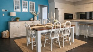 A modern kitchen with white cabinets, a dining area with a wooden table and white chairs, and a blue accent wall. - Coastal retreat 2km from the beach (Golden Bay)