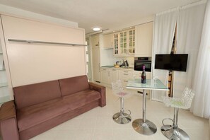 A compact living space with a sofa, a glass dining table, two chairs, a TV, and a kitchenette. - L'ESCALE - Studio Héliopolis D 6 - Luxury - 2 people (AGDE)