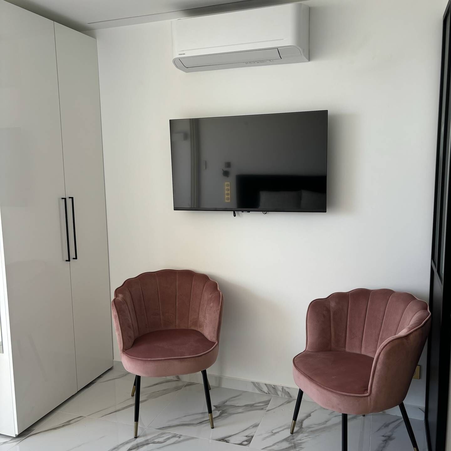 A modern room with a flat-screen TV mounted on the wall, two pink upholstered chairs, a white wardrobe, and a marble floor.