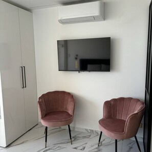 A modern room with a flat-screen TV mounted on the wall, two pink upholstered chairs, a white wardrobe, and a marble floor. - Persuasion - Studio Héliopolis D 8 - Luxe - 2 people (AGDE)