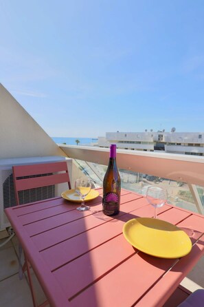Une table rose avec une bouteille de vin et deux verres, placée sur un balcon donnant sur un immeuble et la mer.