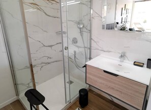 A modern bathroom with a glass shower enclosure, a white sink with a wooden cabinet, and a mirror above the sink. - TROPIC -Studio Héliopolis D 26 - Luxury - 2 people (AGDE)