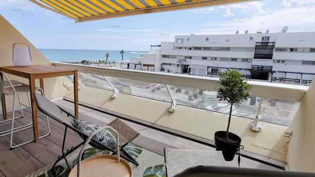 Varanda com mesa de madeira, dois banquetes, uma pequena planta em vaso e vista para a praia e edifícios ao fundo.