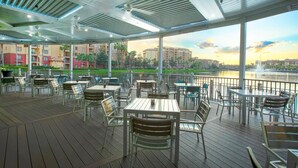 Un patio al aire libre con mesas y sillas, con vistas a un río y edificios al fondo.