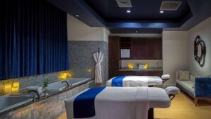 Un cuarto de spa con dos camillas de masaje, un ventanal con cortinas, un lavamanos y una pequeña cocina.